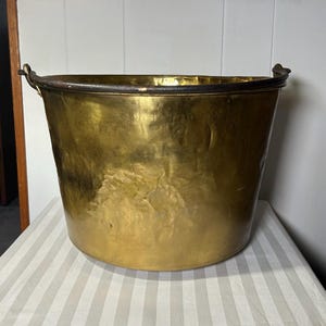 May include: A large, antique brass bucket with a wide, open top and two curved handles. The bucket has a hammered texture and a dark rim. The brass has a warm, golden hue, and the bucket sits on a striped surface.
