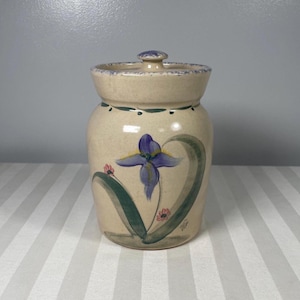 以下が含まれることがあります： 青いアヤメの花と緑の葉を手描きした、蓋付きのベージュ色の陶器の壺。壺は丸みを帯びた形で、蓋には小さなつまみがあります。デザインには小さなピンクの花と緑の縁が含まれています。