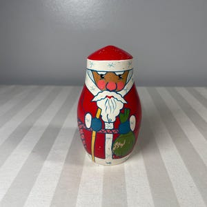 Pode incluir: Uma figura de madeira do Pai Natal com chapéu e casaco vermelhos, barba branca e luvas azuis. O Pai Natal segura um bastão e um saco verde. A figura é pintada com um estilo de arte popular e tem uma forma arredondada.