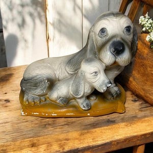 May include: A ceramic figurine of two grey dogs, one larger than the other, sitting on a yellow base. The dogs are positioned as if the smaller dog is resting its head on the larger dog's back.