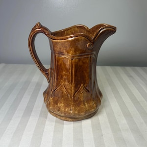 Pode incluir: Um jarro de cerâmica castanho com uma pega e um bico. O jarro tem um design decorativo na frente e uma borda ondulada. O jarro é provavelmente vintage e é adequado para servir bebidas.