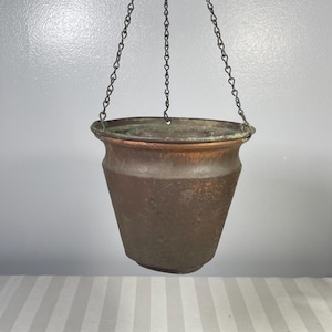 May include: A vintage copper hanging planter with a hammered texture, suspended by a chain. The planter has a flared rim and a dark patina, suggesting age and use. The background is a neutral grey.