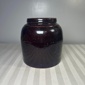 Puede incluir: Un tarro de gres vidriado de color marrón oscuro, con un cuerpo redondeado y un borde ligeramente ensanchado. El tarro tiene un acabado brillante y parece ser vintage. El tarro está sobre una superficie rayada.