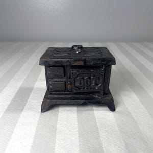 以下が含まれることがあります： 濃い茶色の鋳鉄製のミニチュアストーブ。ストーブは長方形で、平らな上面、小さな煙突、装飾的なディテールがあります。前面には「DOT」の文字が見えます。