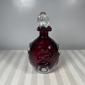 May include: A ruby red glass perfume bottle with a clear glass stopper. The bottle has a bulbous body with a textured design and a ruffled neck. The stopper is elongated and has a rounded top.