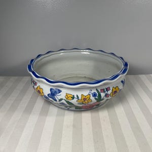 May include: A white ceramic bowl with a scalloped rim and a blue border. The bowl is decorated with colorful floral and butterfly designs. The bowl is likely used for decorative purposes or as a planter.