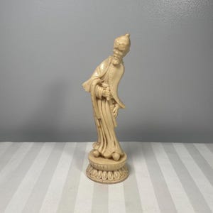 May include: Ivory-colored figurine of a man in traditional Asian attire. The figure has a long beard and is wearing a robe with long sleeves. The figurine is standing on a decorative base with a floral design. The background is a light gray wall.