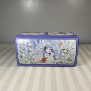 May include: A rectangular, lilac tin box with a winter scene featuring snowmen and snow. The box has a hinged lid with a rounded edge. The main image shows a snowman with a hat and scarf, surrounded by smaller snowmen.