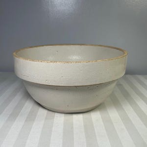 May include: A cream-coloured ceramic mixing bowl with a wide rim and a textured surface. The bowl has a classic, rounded shape and a light brown rim. The bowl is likely vintage or antique.