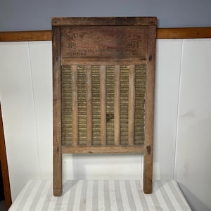 Puede incluir: Un antiguo lavadero de madera con marco rectangular y superficie de fregado texturizada. La sección superior presenta texto estampado, incluyendo "MEMPHIS" y "CHICAGO". El lavadero tiene un aspecto rústico y envejecido.