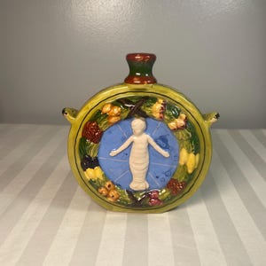 May include: A round, ceramic flask with a yellow border and a blue center featuring a white figure. The flask has a green and red stopper and small handles. The border is decorated with colorful floral and fruit motifs.