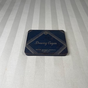 May include: A vintage, rectangular, blue metal box of drawing crayons. The box features the words "Drawing Crayons" in a decorative font, with the brand name "Milton Bradley Company" below. The box has a geometric design and is made in the USA.