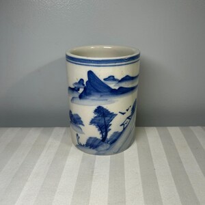 Puede incluir: Una taza cilíndrica de cerámica con un diseño de paisaje azul y blanco. El diseño presenta montañas, árboles y una pequeña casa. La taza tiene un interior blanco y un borde azul.