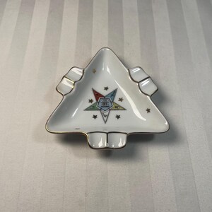 Puede incluir: Cenicero triangular blanco con ribete dorado. El cenicero presenta un emblema colorido con estrellas y las letras "OES". El cenicero tiene un diseño pequeño y poco profundo con un acabado liso y brillante. El cenicero es probablemente de cerámica o porcelana.