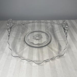 May include: Clear glass serving dish with a scalloped edge and two ornate handles. The center of the dish features a raised floral design. The dish is square with rounded corners and is set against a striped background.