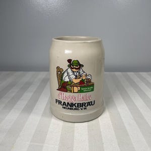 May include: A beige ceramic beer stein with a cartoon illustration of a man in a green hat and a beer. The stein has the text "d'Hogga Halbe FRANKBRAU NEUNBURG V.W."