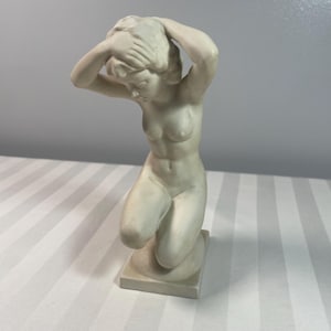 Puede incluir: Figura arrodillada de color crema de una persona con las manos en la cabeza. La escultura está sobre una base cuadrada y tiene un acabado liso y mate. La figura es desnuda y los detalles están finamente esculpidos.