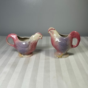 Puede incluir: Dos jarras de crema de cerámica con forma de gallo. Las jarras están pintadas con un degradado de colores, incluyendo rosa, morado y blanco. Cada jarra tiene un asa y un pico. Las jarras con forma de gallo son decorativas y funcionales.