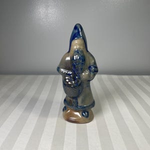 May include: A ceramic figurine of a hooded figure, possibly a gnome or Santa Claus, with a blue hat and trim. The figure is holding a sack or basket. The color palette includes blue, brown, and beige tones.