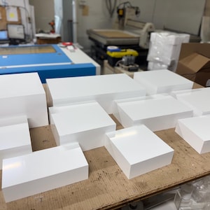 May include: A collection of white acrylic display risers of various sizes, arranged on a wooden surface. The risers are ideal for showcasing products or displaying merchandise.