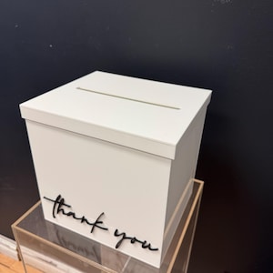 May include: A white wooden box with a slot on top, resting on a clear acrylic stand. The words "thank you" are written in black script on the front of the box. The background is a dark navy blue wall.