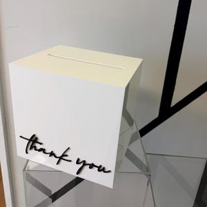 Peut inclure: Une boîte carrée blanche avec une fente horizontale sur la surface supérieure. La face avant présente les mots thank you en lettres cursives noires en relief. La boîte est construite avec une finition lisse et un design géométrique.
