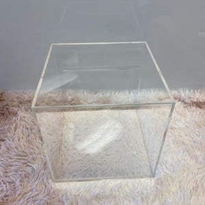 May include: A transparent acrylic box with a slot on the top. The box is a cube shape and is placed on a fluffy, white surface. The clear design allows for a view of the inside.