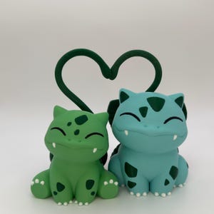May include: Two smiling, cartoon-like figures, one green and one blue, sit side-by-side. They have dark green spots and small white teeth. A dark green, heart-shaped vine arches above them. The figures are on a white surface.