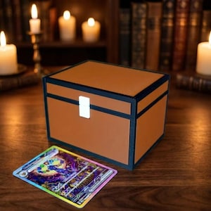 Könnte beinhalten: Eine rechteckige braune Aufbewahrungsbox mit schwarzem Rand und einem weißen Verschluss, begleitet von einer einzelnen rechteckigen Sammelkarte mit einer farbenfrohen, detaillierten grafischen Illustration in Violett-, Blau- und Goldtönen.