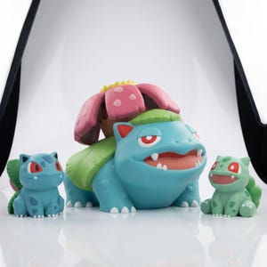 May include: A set of three vinyl figures featuring a large central creature with a pink flower on its back, flanked by two smaller creatures. The central figure is light blue with a green leaf base, while the smaller figures are blue and green.