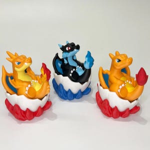 May include: Three colorful dragon figurines in eggshell containers. The dragons are orange, black, and blue, with flames and wings. The eggshells are white with red and blue accents. The figurines are on a white surface.