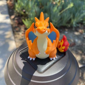 May include: A 3D-printed orange and cream-colored Charizard figure with blue wings and a fiery tail. The figure is perched on a black square base, set against a blurred outdoor background.