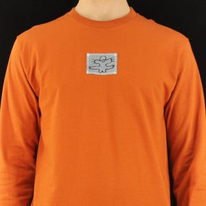 May include: A long-sleeve crew neck shirt in a solid burnt orange color. Centered on the chest is a small, rectangular light gray patch featuring a black line drawing of a stylized creature with outstretched limbs and a round head.