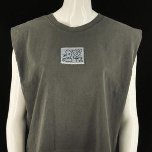 May include: A charcoal gray sleeveless t-shirt with a crew neckline and padded shoulders. The shirt features a small, rectangular, light-colored patch on the center chest with a black line drawing of two abstract figures.