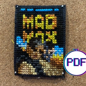 May include: A black and brown cross-stitch pattern with the words "MOD VTH" in yellow thread. The pattern is framed and pinned to a corkboard.