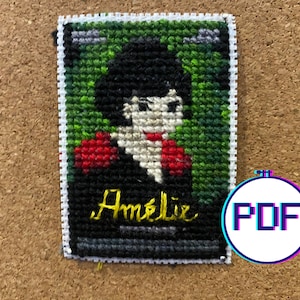 May include: Cross-stitch pattern of a woman with black hair and a red shirt. The pattern is framed in a white border and the name "Amelie" is stitched in yellow below the image.