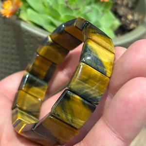 Op de afbeelding: Een armband bestaande uit rechthoekige tijgeroog stenen kralen. De kralen hebben een gepolijste afwerking met afwisselende banden van goudgeel en donkerbruin, wat een chatoyant effect creëert over het oppervlak van elk individueel steensegment.