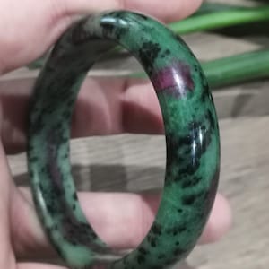 Op de afbeelding: Een gepolijste jade armband. De armband heeft een diepgroene kleur met zwarte en rode insluitsels. Het gladde, cirkelvormige ontwerp wordt in een hand gehouden en toont de natuurlijke variaties in de steen.