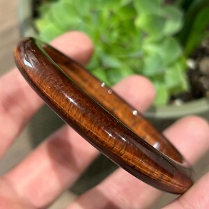 Peut inclure: Un bracelet jonc circulaire poli, fabriqué à partir d'un bois brun foncé riche avec une finition lisse et brillante. Le grain du bois présente des stries verticales visibles et des motifs naturels sur toute la surface de l'anneau solide.