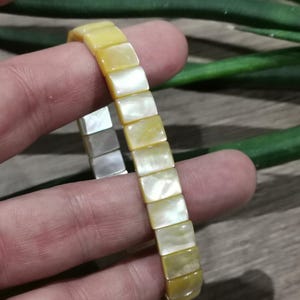 May include: A yellow and white bracelet composed of rectangular, iridescent beads. The bracelet is held by a hand, highlighting its colour and design. The beads have a pearlescent sheen, reflecting light.