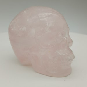 Peut inclure: Un crâne de cristal rose pâle sculpté dans du quartz rose. Le crâne est lisse et poli, avec une apparence translucide.