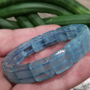Puede incluir: Un brazalete de aguamarina azul claro compuesto por cuentas rectangulares. El brazalete se sostiene en una mano, con un fondo borroso de objetos verdes y blancos. Las cuentas son translúcidas y tienen un sutil tono azul.