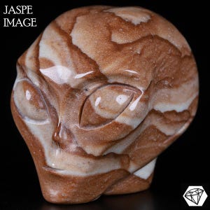 Peut inclure: Une sculpture de tête d'extraterrestre en jaspe marron et blanc. La sculpture a de grands yeux ovales et une surface lisse et polie. Le texte "JASPE IMAGE" est dans le coin supérieur gauche.