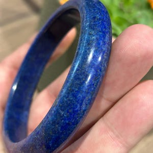 Może przedstawiać: Polerowana bransoletka typu bangle wykonana z ciemnoniebieskiego kamienia lapis lazuli. Okrągła obręcz ma gładką, zaokrągloną powierzchnię z naturalnymi wariacjami kolorystycznymi, w tym drobinkami złotego pirytu wtopionymi w ciemnoniebieski minerał.