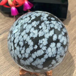 Peut inclure: Une sphère en obsidienne flocon de neige noire, d'environ 10 cm de diamètre, avec un motif unique d'inclusions blanches en forme de flocons de neige. La sphère repose sur un petit support en bois, avec un objet rectangulaire noir et des fleurs rouges en arrière-plan.