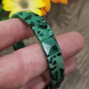 Può includere: Un braccialetto di pietra verde e nera. Il braccialetto è composto da pietre rettangolari e sfaccettate. Le pietre sono di un colore verde intenso con inclusioni nere. Il braccialetto è tenuto davanti a uno sfondo sfocato di fiori gialli.
