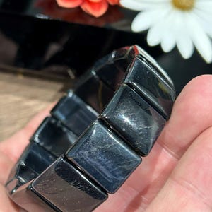 Peut inclure: Un bracelet noir composé de pierres rectangulaires polies. Le bracelet est tenu dans une main, avec un arrière-plan flou avec des fleurs. Les pierres ont une qualité réfléchissante, captant la lumière et affichant de subtiles variations de couleur.