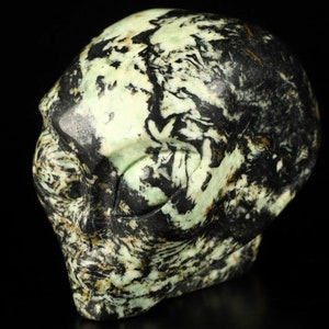 Peut inclure: Un crâne d'alien vert et noir sculpté dans une pierre avec un motif tourbillonnant. Le crâne est détaillé avec des yeux, un nez et une bouche.