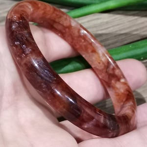 Peut inclure: Un bracelet jonc en pierre précieuse polie, de couleur brun rougeâtre. La pierre translucide présente un mélange de tons foncés et clairs, avec une forme lisse et arrondie. Le bracelet est tenu dans une main.