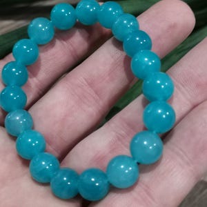 Peut inclure: Un bracelet de perles turquoise. Le bracelet est composé de perles rondes et lisses d'un bleu-vert vif. Les perles sont enfilées pour former un cercle complet. Le bracelet est présenté sur une main.